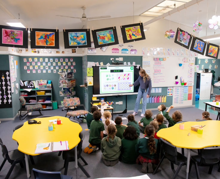 Year 3/4 classroom. Bright colourful artworks around the room. Students sitting on the flooer while teacher uses a Smartboard for their lesson.