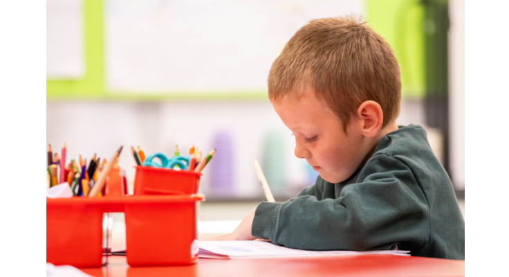 A Kindergarten boy sitting at his desk completing some writing. He has a tin of pencils on his desk also.